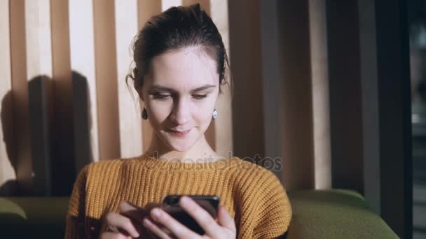 Retrato de una joven hermosa mujer sentada en un sofá y usando un teléfono inteligente. Chica charlando con amigos en la mañana . — Vídeo de stock