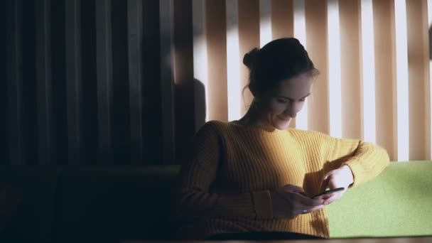 Porträtt av unga leende kvinna sitter i soffa och med smartphone. Flicka surfa på Internet på morgonen — Stockvideo