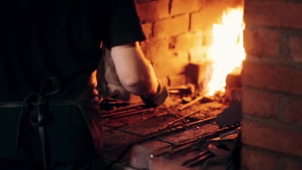 Schmied in Schürze am Arbeitsplatz. Nahaufnahme eines Mannes, der das heiße Metall mit Feuer aus dem Ofen holt. — Stockvideo