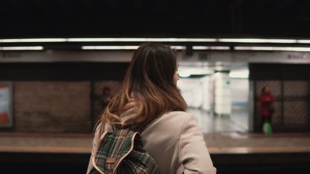 Mujer morena de pie en la estación de tren y esperando su tren. Chica en metro por la noche se ve en tranvías rápidos — Vídeo de stock