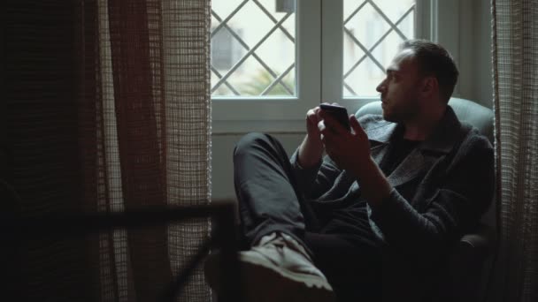 Stylish man sitting in armchair and looking at window. Young attractive male uses smartphone in living room. — Stock Video