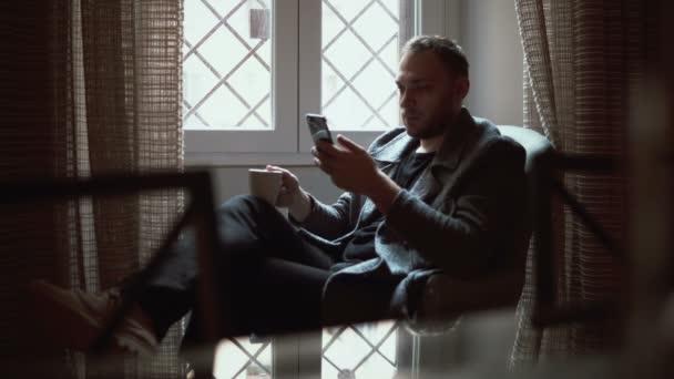 Retrato de homem sentado em poltrona na sala de estar e usando smartphone. Masculino navegar na Internet e bebe café . — Vídeo de Stock
