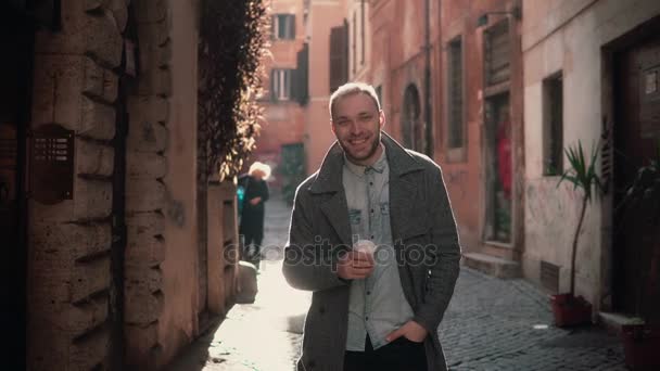 Retrato de un joven guapo mirando a la cámara. Feliz hombre sonriente de pie en la calle de la mañana, bebiendo café . — Vídeo de stock