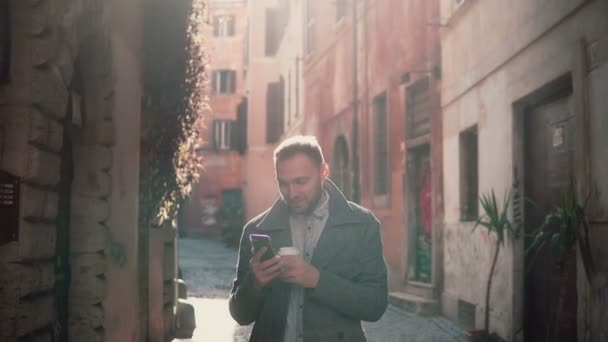 Χαμογελαστός νεαρός περπατώντας στην πόλη τη πρωί και χρησιμοποιώντας το smartphone. Τύπος περιηγηθείτε στο διαδίκτυο, texting με κάποιο. — Αρχείο Βίντεο