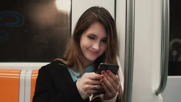 Retrato de menina atraente no trem de metrô usando smartphone. Jovem conversando com amigos e sorrindo . — Fotografia de Stock