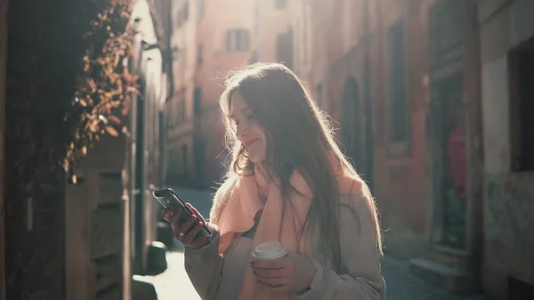 Retrato de jovem mulher feliz andando na cidade e usando smartphone. Menina navegar na Internet e beber café . — Fotografia de Stock