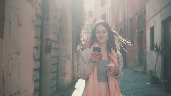 Jovem mulher feliz andando no centro da cidade de manhã, usando smartphone. Menina navegar na Internet, mensagens de texto com alguém . — Fotografia de Stock