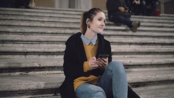 Chica estudiante sentada en la noche en la escalera y mirando a alguien, sonrisa. Mujer utilizar el teléfono inteligente, navegar por Internet . — Vídeos de Stock