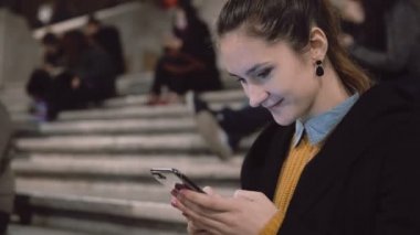 Kalabalığın içinde merdiven üzerinde oturan ve smartphone kullanarak genç esmer kadın. Etrafa ve gülümseyen kız.