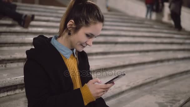 Retrato de una joven sentada en la escalera y sosteniendo un smartphone. Chica utiliza la pantalla táctil para navegar por Internet . — Vídeos de Stock