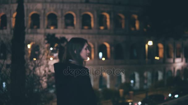 Retrato de una joven morena de pie cerca del Coliseo en Roma, Italia por la noche. Chica se gira y mira a la cámara . — Vídeo de stock