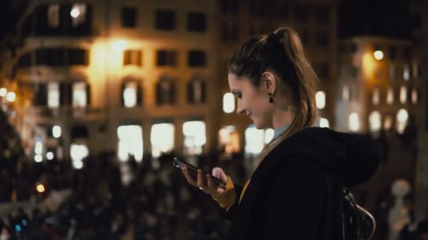 Šťastná mladá žena stojící v davu a pomocí smartphonu. Dívka procházky a chatování s přáteli večer. — Stock video