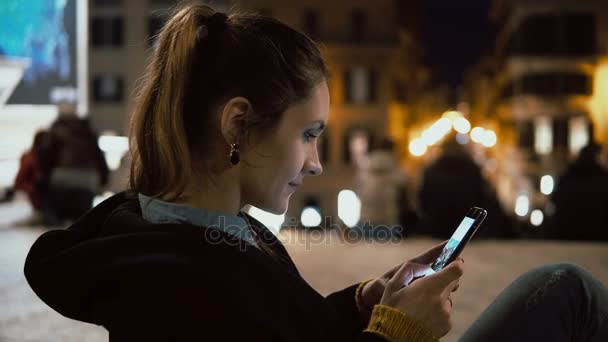 Attraktiv brunett kvinna sitter i centrum i kväll och ser foto på smartphone med pekskärm teknik. — Stockvideo