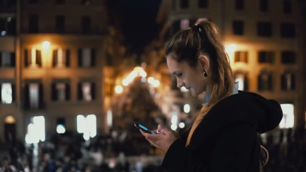 Молода приваблива жінка стоїть в центрі міста ввечері і користується смартфоном. Натовп і вогні на фоні — стокове відео
