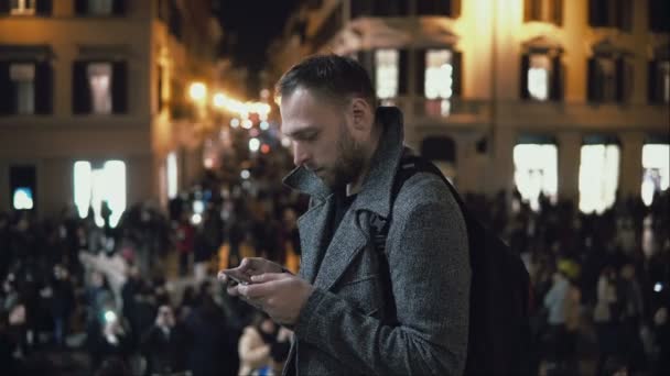 Joven turista guapo de pie en la multitud por la noche. Macho utilizar el teléfono inteligente para encontrar la dirección . — Vídeo de stock