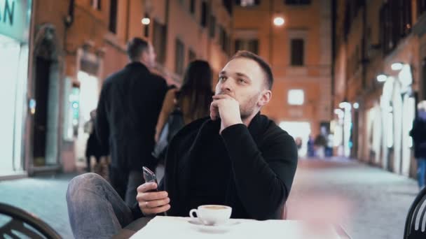 Joven hombre guapo sentado afuera en la cafetería de la ciudad y sosteniendo el teléfono inteligente. Hombre tomando café por la noche . — Vídeos de Stock