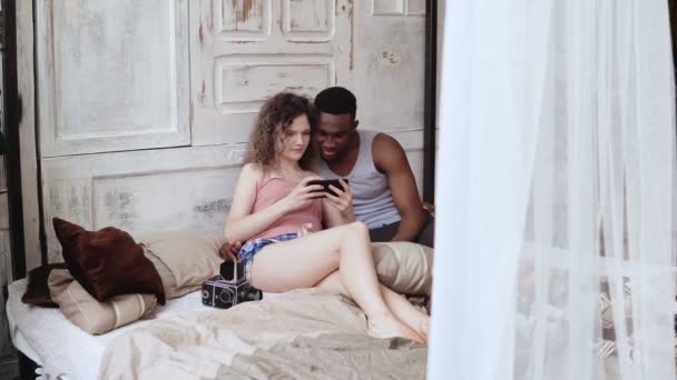 Homem e mulher de pijama sentados na cama. Casal multirracial tira foto selfie, usar smartphone e rindo . — Vídeo de Stock