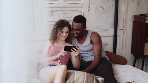 Многорасовая пара в пижаме сидит на кровати и пользуется смартфоном. Африканский мужчина и европейская женщина в Интернете, улыбаются — стоковое видео