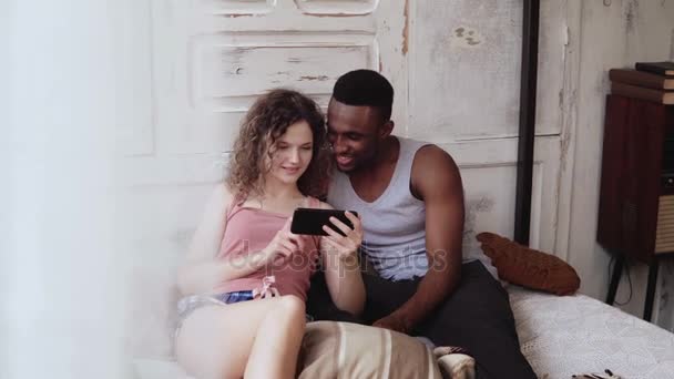 Multiraciale paar in pyjama zittend op bed en gebruik smartphone. Afrikaanse man en Europese vrouw surfen op het Internet, smile — Stockvideo