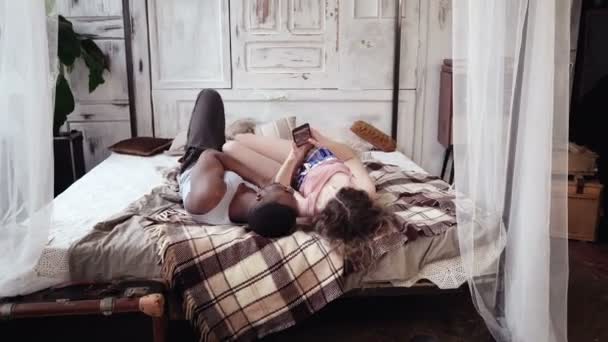 Junges multiethnisches Paar im Schlafanzug, auf dem Bett liegend und Fotos schauend. Mann und Frau nutzen Smartphone mit Touchscreen. — Stockvideo