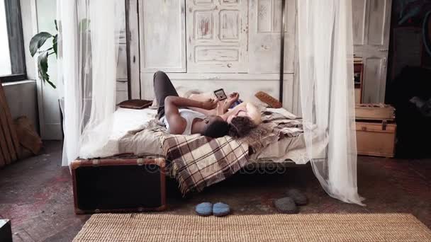 Çok ırklı çift yatakta yatıyordu görünümünü arka ve akıllı telefon kullanın. Afrika Avrupa kadın ve görünüyor fotoğraf. — Stok video