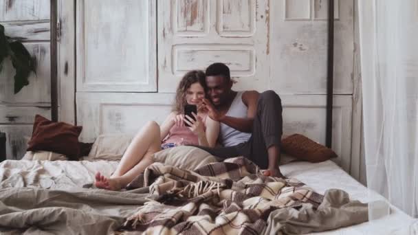 Ein Paar im Schlafanzug sitzt auf dem Bett und nutzt das Smartphone. Afrikaner und Europäerin suchen Fotos und lachen — Stockvideo