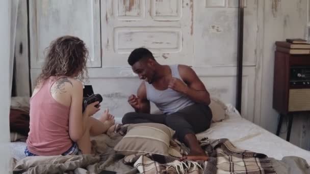 Photoshoot multiracial. Photographe femme prenant une photo de l'homme drôle en pyjama sur la caméra du film en mouvement sur le lit . — Video