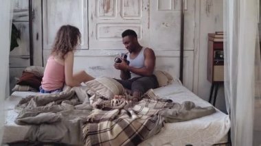Genç adam alarak hareket-ince tabaka fotoğraf makinesi üzerinde pijama kadın fotoğrafı. Çok ırklı çift yatağa sabah oturan.