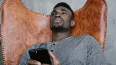 Afrika erkek sandalyede oturan ve düşünme, akıllı telefon tutarak. Adam iyi bir şey hatırlar ve mesajlar yazarak başlar.