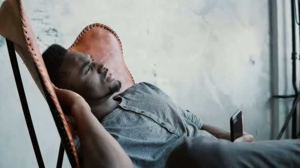 Porträt eines jungen Afrikaners im Stuhl. Mann überlegt ernsthaft und hält Smartphone in der Hand. Typ tippt Nachricht ein, lächelt. — Stockvideo