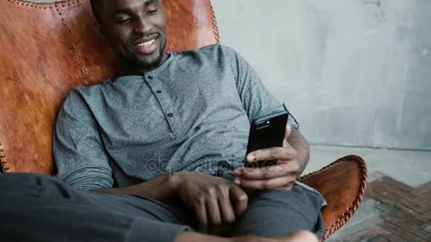 Retrato del hombre africano sentado en silla, usando Smartphone. El hombre lee mensajes y se ríe. Chico escribiendo masajes, sonriendo — Vídeo de stock