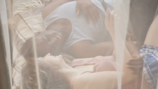 침대에 아름 다운 multiracial 커플입니다. 남자와 여자 잠 옷 사기에 사용 스마트폰, 웃음. 커튼을 통해 볼. — 비디오