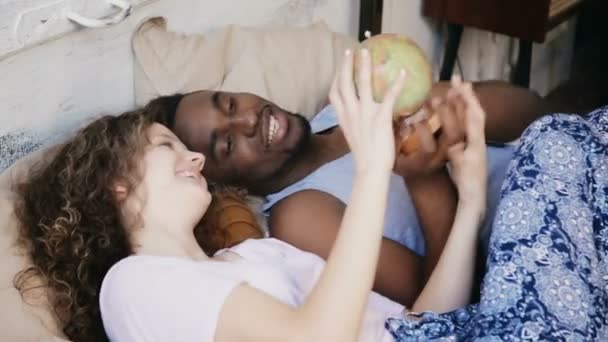 Feliz casal multirracial deitado na cama e torcer o globo. Homem e mulher aponta o destino para viajar, rir . — Vídeo de Stock