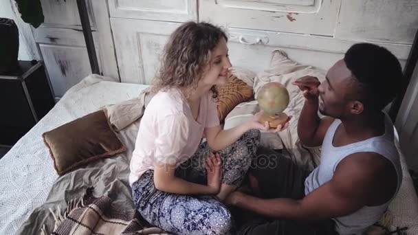 Woman twists the globe, man picks the location to traveling. Multiracial couple in pajamas rejoice, hug and kiss. — Stock Video
