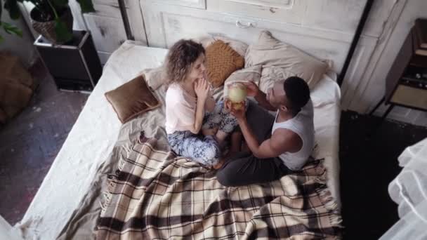 Casal multiétnico sentar-se na cama e torcer o globo. Homem e mulher escolhem o destino para viajar, abraçar e beijar . — Vídeo de Stock