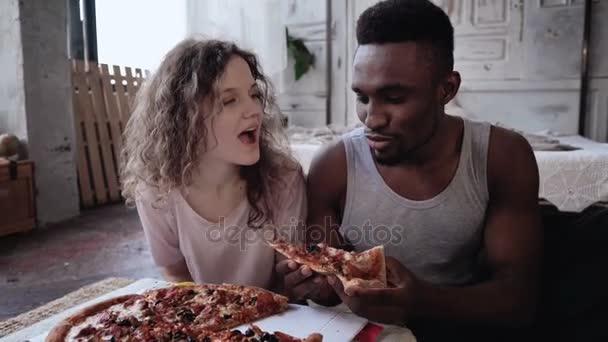 Feliz casal multiétnico de pijama comendo fast food. Mulher faminta espera a refeição, homem alimenta-lhe uma fatia de pizza . — Vídeo de Stock