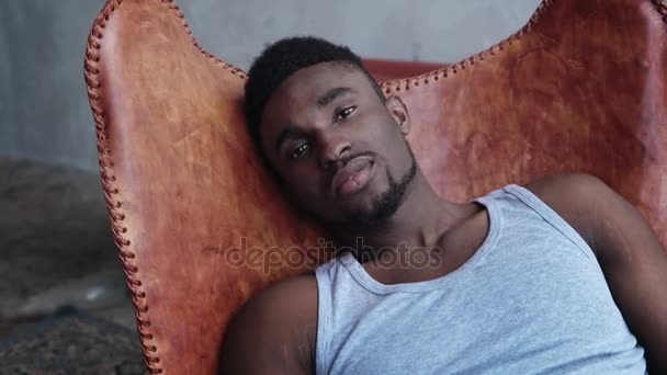 Porträt eines jungen gut aussehenden afrikanischen Mannes, der auf dem Ledersessel sitzt und in die Kamera blickt. seriöses Modell männlich. — Stockvideo