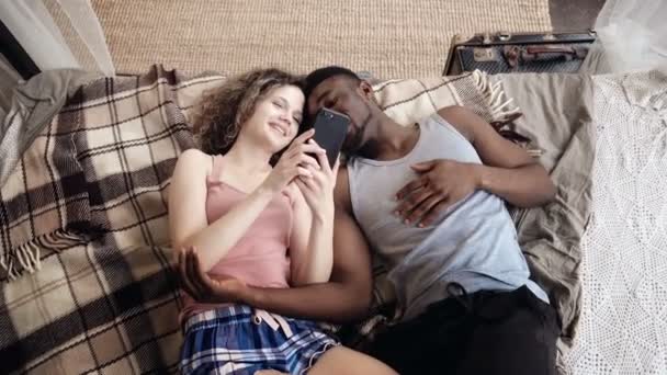 Multiracial couple lying on bed. Man and woman spend time together with joy and fan, use Smartphone, surf the internet. — Stock Video