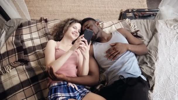 Mnohonárodnostní pár ležící na posteli. Muž a žena trávit čas s radostí a ventilátor, použijte Smartphone, surfovat po internetu.