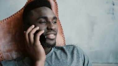 Sandalyede oturan ve Smartphone'da konuşan genç Afrika erkek portresi. Adam gülüyor ve onun baş kabul sallıyor.