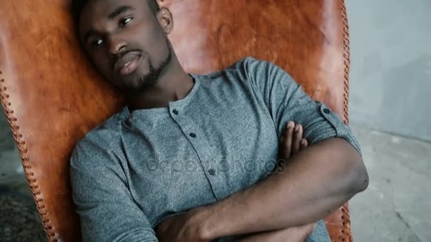 Hombre africano guapo con la barba está sentado en la silla y sonriendo. Un hombre se ve soñador, reflexivo y tranquilo . — Vídeos de Stock