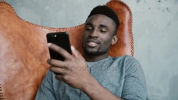 Retrato de un joven africano sentado en la silla, usando Smartphone. Un guapo macho sonriendo y hablando por teléfono . — Vídeos de Stock