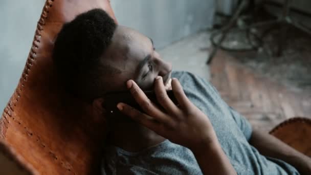 Retrato de un hombre africano sentado en una silla en una habitación luminosa y hablando en el Smartphone. Hombre riendo y divirtiéndose . — Vídeos de Stock