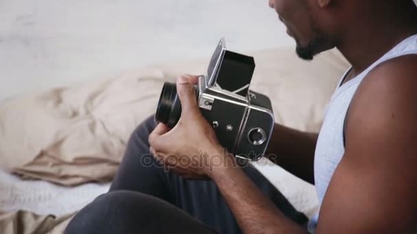 Jovem africano de pijama sentado na cama e segurando a velha fotocâmera. Cara falando foto na câmera de filme em movimento . — Vídeo de Stock