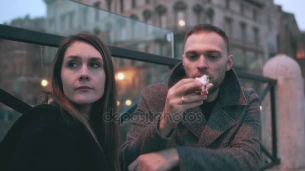 Молодая женщина сидит снаружи, мужчина приходит и садится рядом с ней. Привлекательная пара ест сэндвичи и разговаривает . — стоковое видео