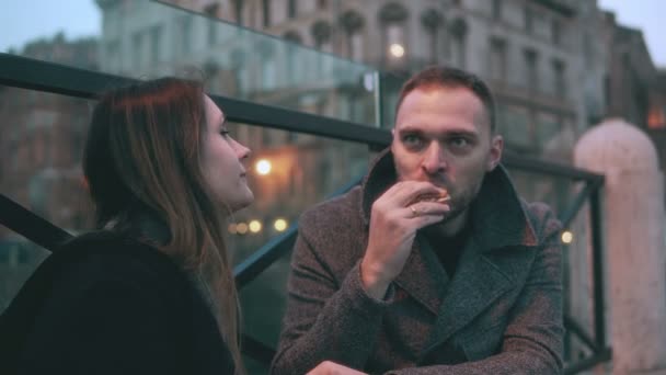 Jovem e mulher, dois amigos sentados na ponte à noite e comendo sanduíches. Casal ter uma pausa . — Vídeo de Stock