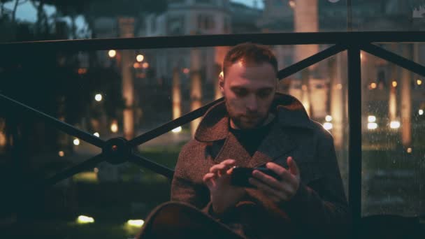 Junger gutaussehender Mann mit Smartphone, Abendstadt im Hintergrund. Mann surft mit Touchscreen-Technologie im Internet. — Stockvideo