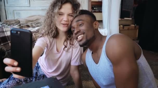 Hermosa pareja multiétnica tomar la foto selfie en el teléfono inteligente. La mujer sostiene el teléfono inteligente, el hombre la besa y se ríe . — Vídeo de stock