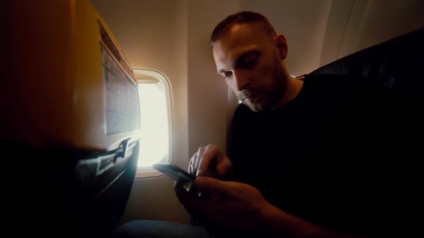 Jovem homem atraente usa smartphone dentro do avião ao lado da janela. Bonito macho viajando pelo ar . — Vídeo de Stock