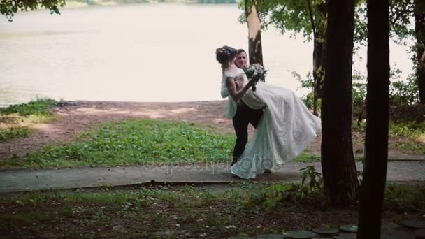 Handsome groom holds his bride in his arms and swirls. Beautiful wedding on a river bank in summertime. — Stock Video